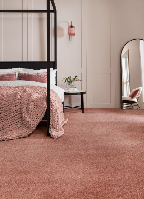 A black bed frame in front of a beige panelled wall. Pink bedding and carpeted floor.