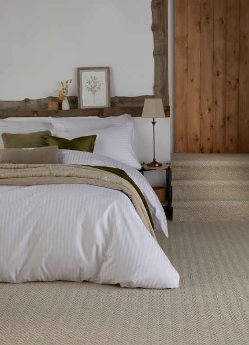 A bedroom with large bed with beige & green tonned throws & cushions. Beige herringbone carpet.